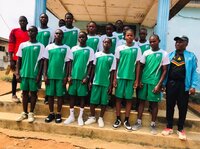 Eine Gruppe junger Fußballspieler in grünen Trikots steht gemeinsam mit einem Trainer vor einem Gebäude in Kamerun und posiert für ein Mannschaftsfoto.