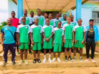 Eine Gruppe junger Fußballspieler in grünen Trikots steht gemeinsam mit einem Trainer vor einem Gebäude in Kamerun und posiert für ein Mannschaftsfoto.